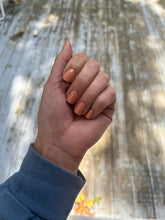 Load image into Gallery viewer, Hand with neutral orange nail polish against a blurred natural background