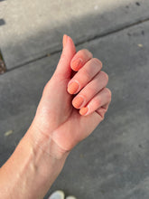 Load image into Gallery viewer, Hand with orange nail polish against a gray pavement background