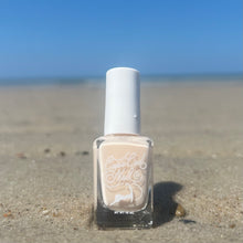 Load image into Gallery viewer, A bottle of beige-colored nail polish displayed on a sandy beach with the ocean in the background.
