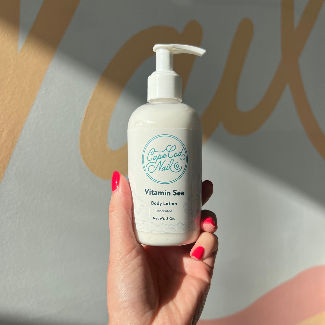 A hand holding a pump bottle of Vitamin Sea body lotion with a label featuring Cape Cod Nail Co. and a beach-themed design.